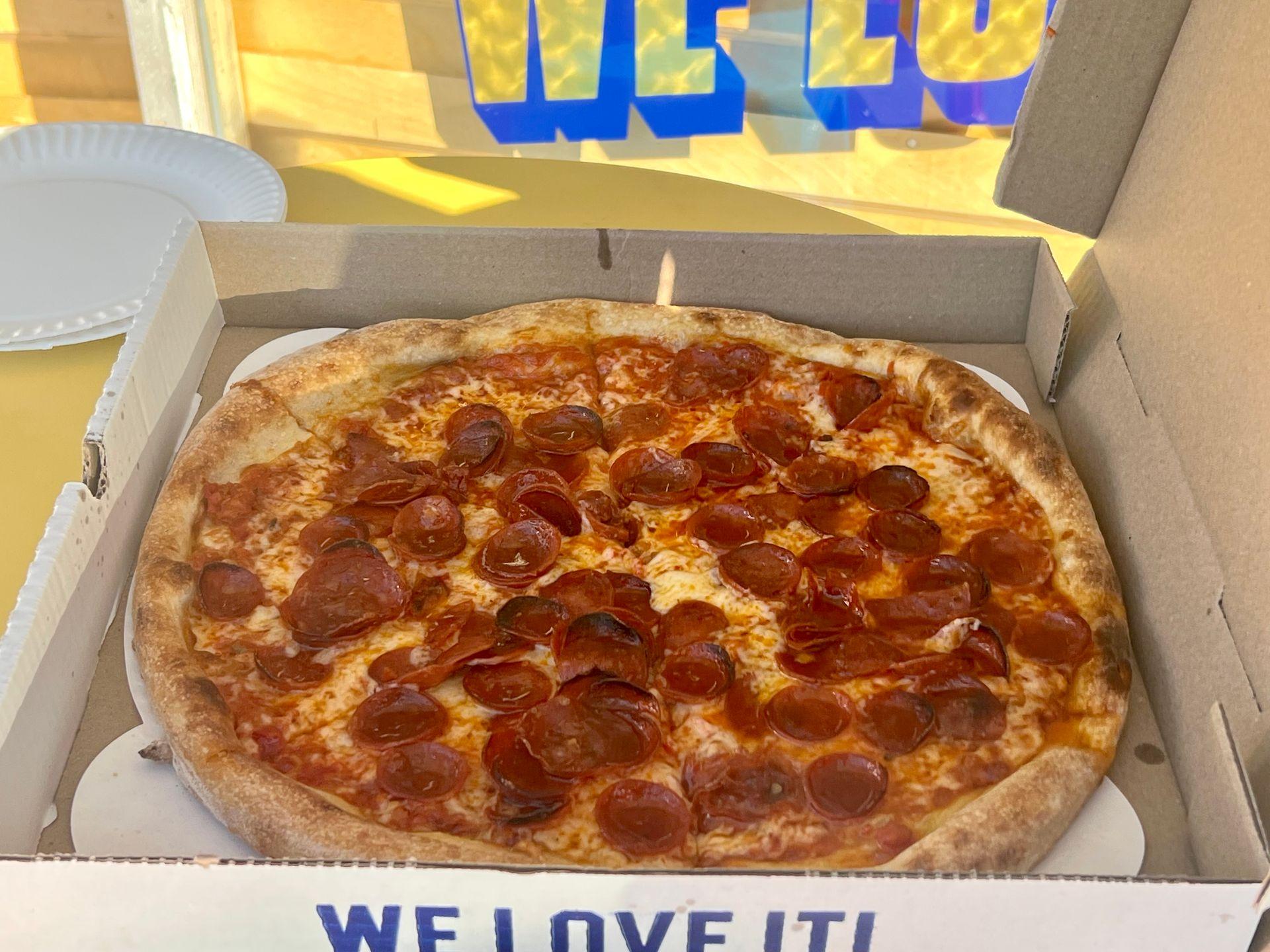 Pepperoni pizza in an open cardboard box on a yellow table with paper plates nearby.