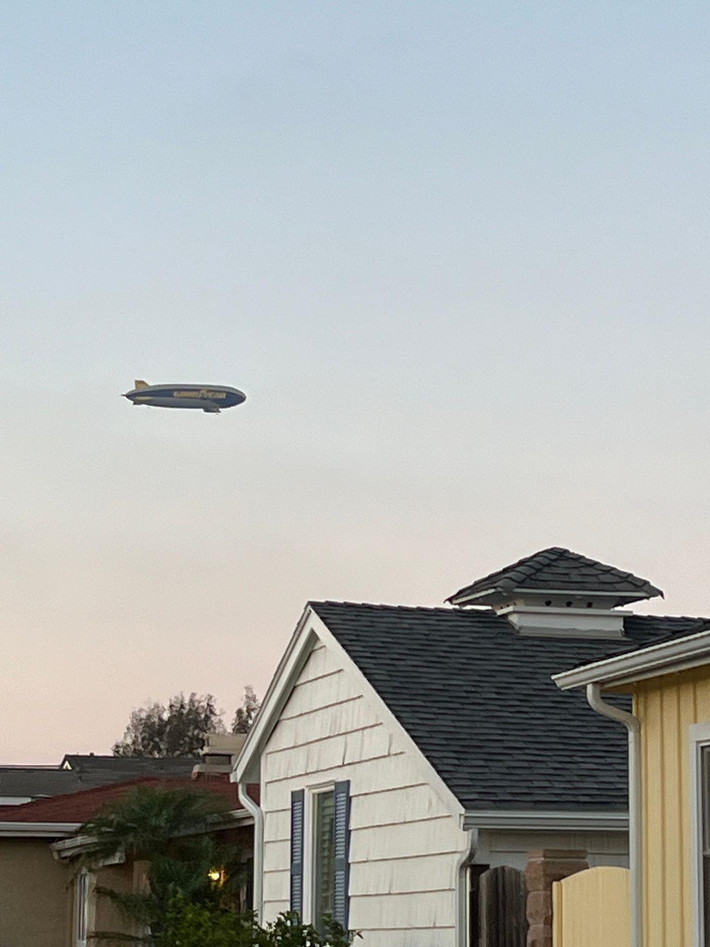 Blimp flying over residential houses at dusk, with a clear sky in the background.