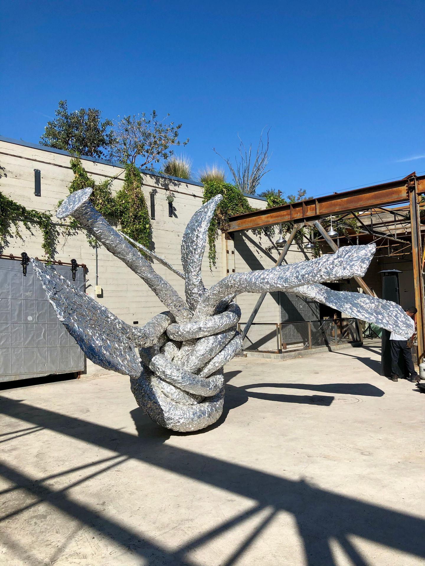Large metallic abstract sculpture with twisted elements situated outdoors against a concrete wall covered in greenery.
