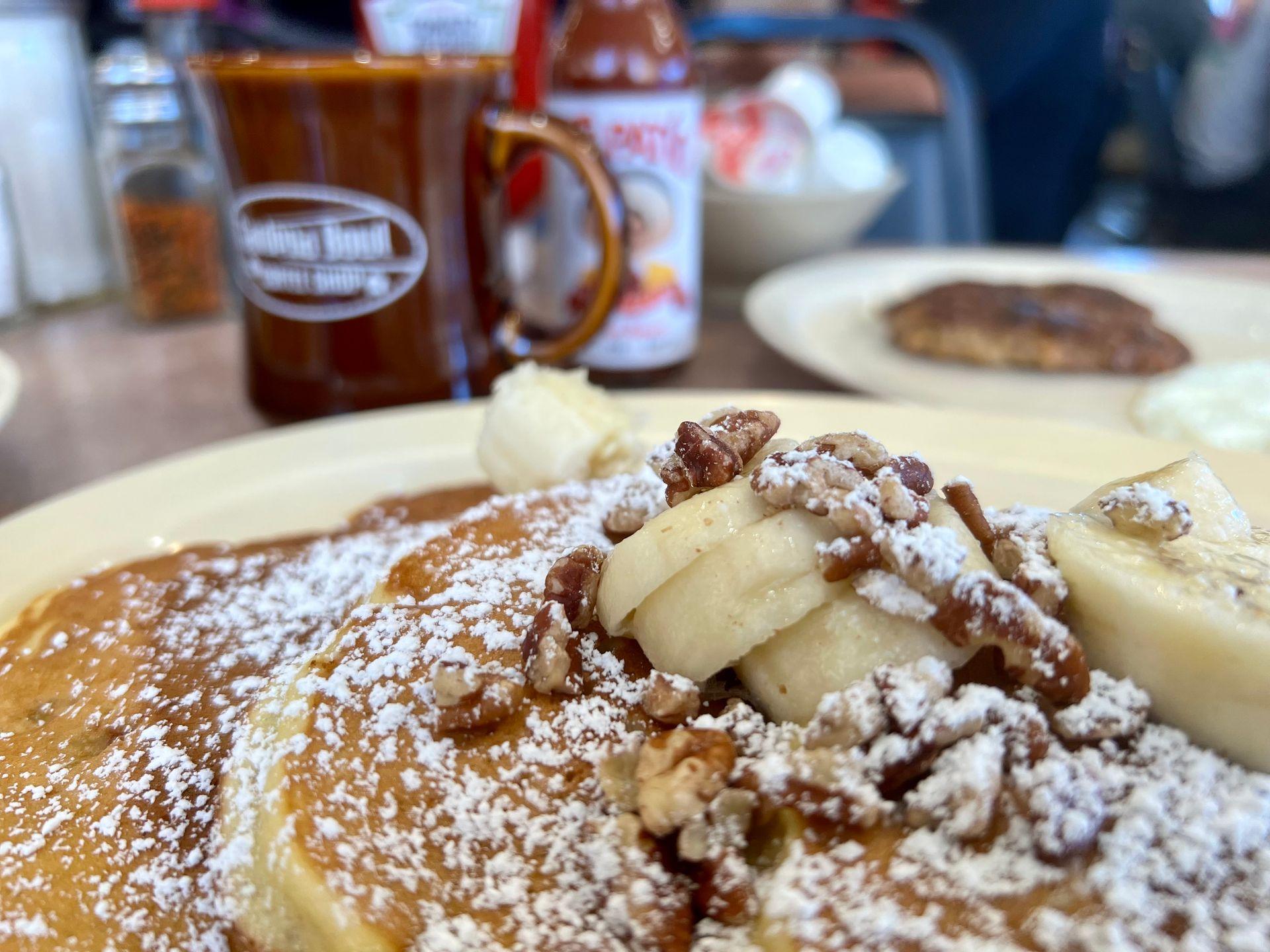 Close-up of pancakes topped with bananas, pecans, and powdered sugar on a plate.