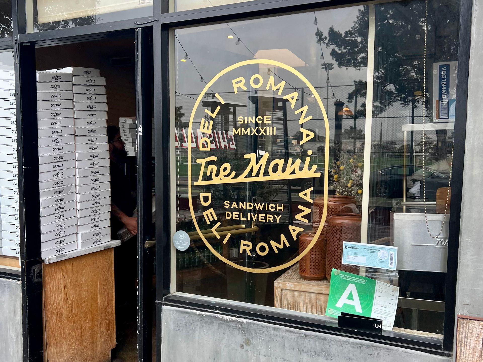 Entrance of deli shop 'Tre Mani' with glass door and stacked boxes inside.