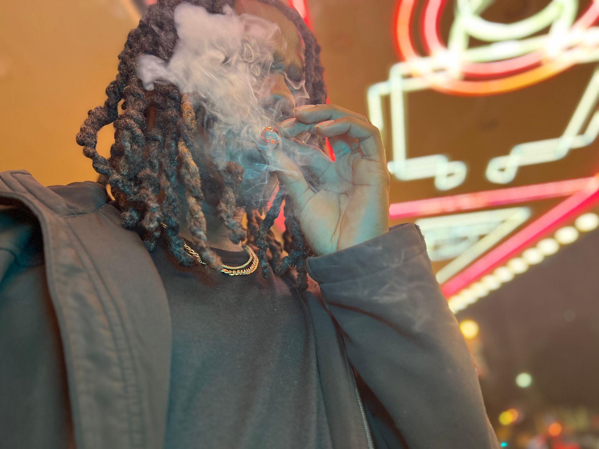 Person with braided hair smoking, neon lights in the background creating a vibrant glow.