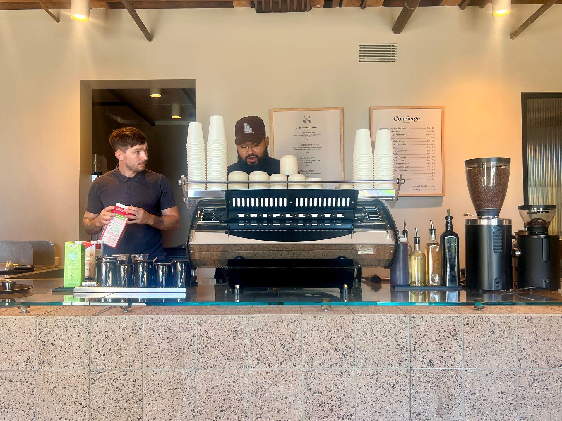 Two people working behind a coffee counter, with cups and coffee-making equipment visible.