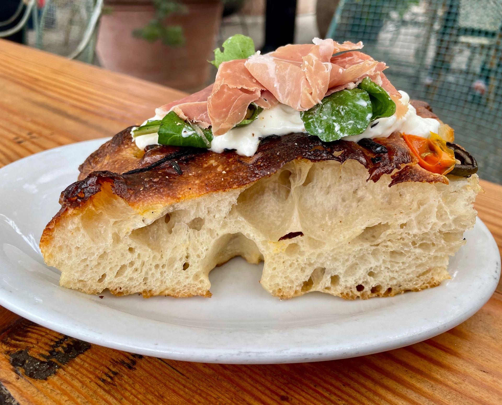 Slice of focaccia topped with prosciutto, greens, and cheese on a white plate.