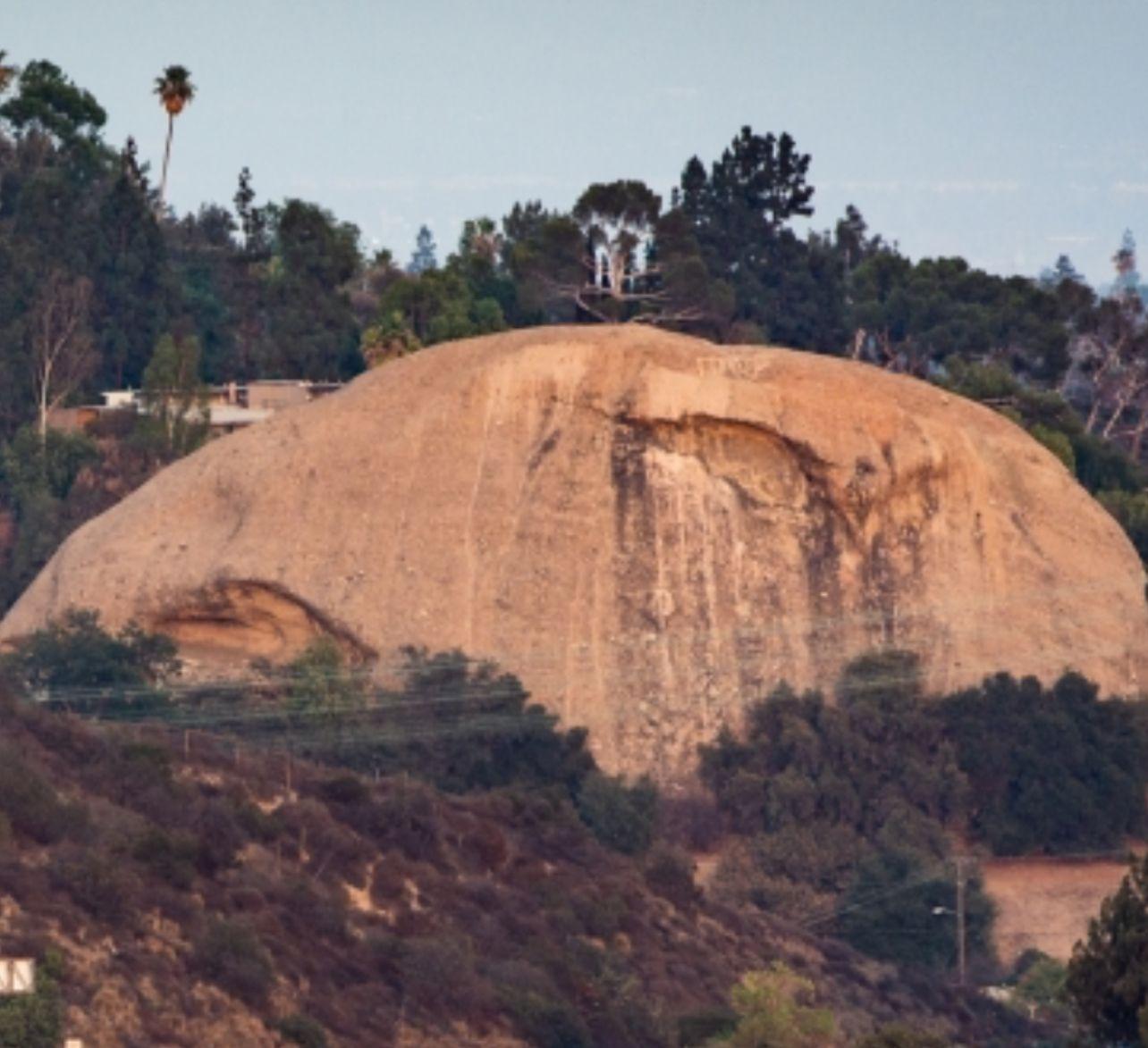 Large rocky hill surrounded by dense forest and vegetation under a clear sky.