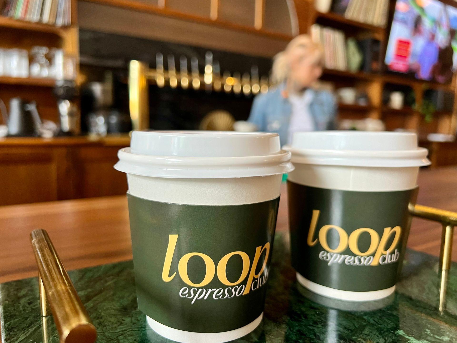 Two coffee cups labeled 'loop espresso club' on a counter with blurred background of cafe interior.