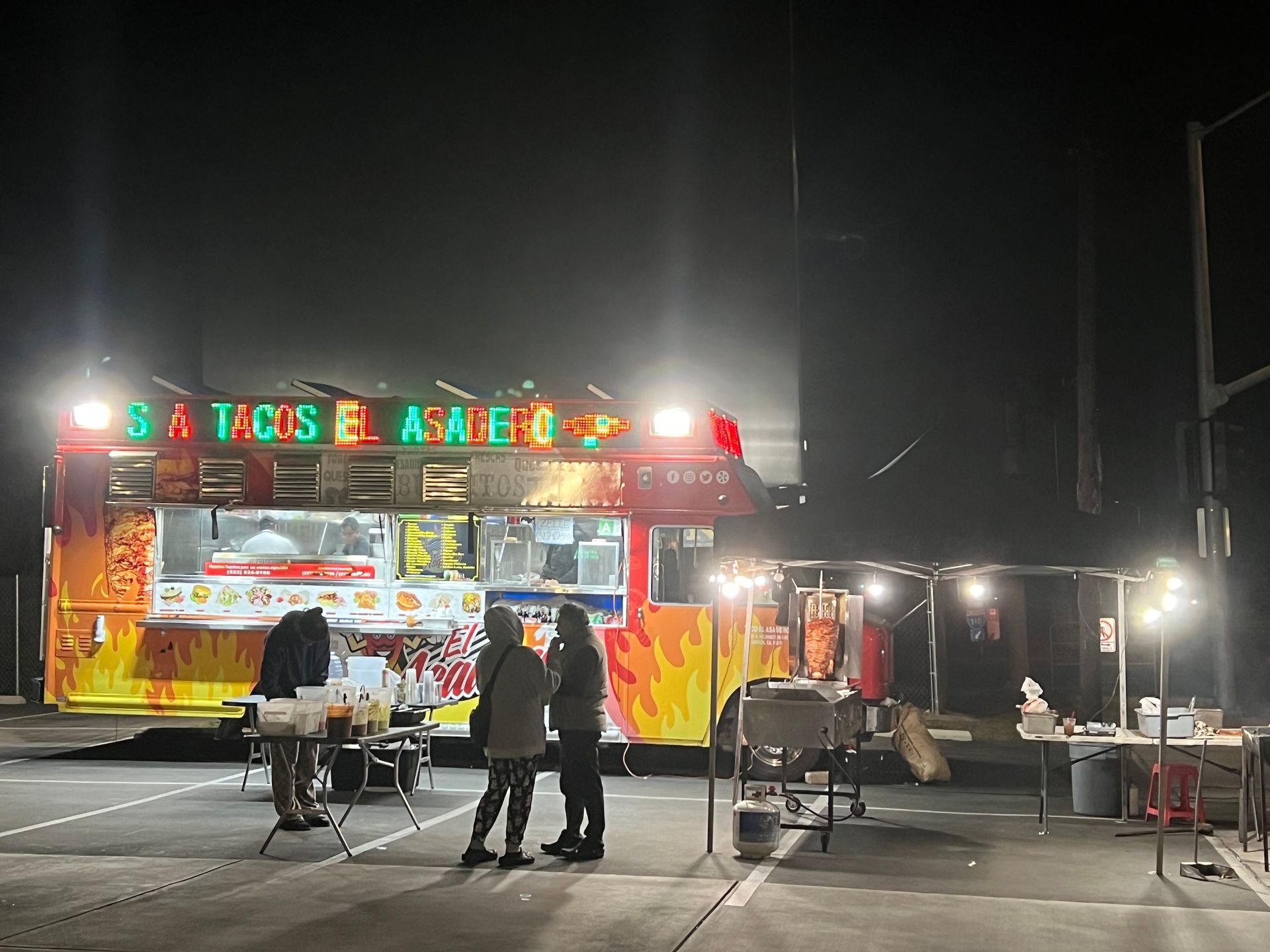 People gathered around a brightly lit taco truck at night, with food preparation visible in the background.