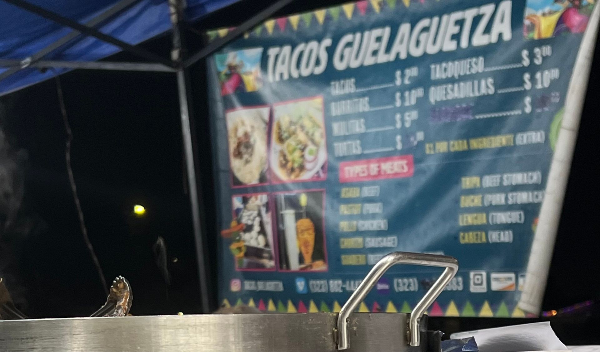 Taco stand menu at night with pricing and options displayed under a blue canopy.