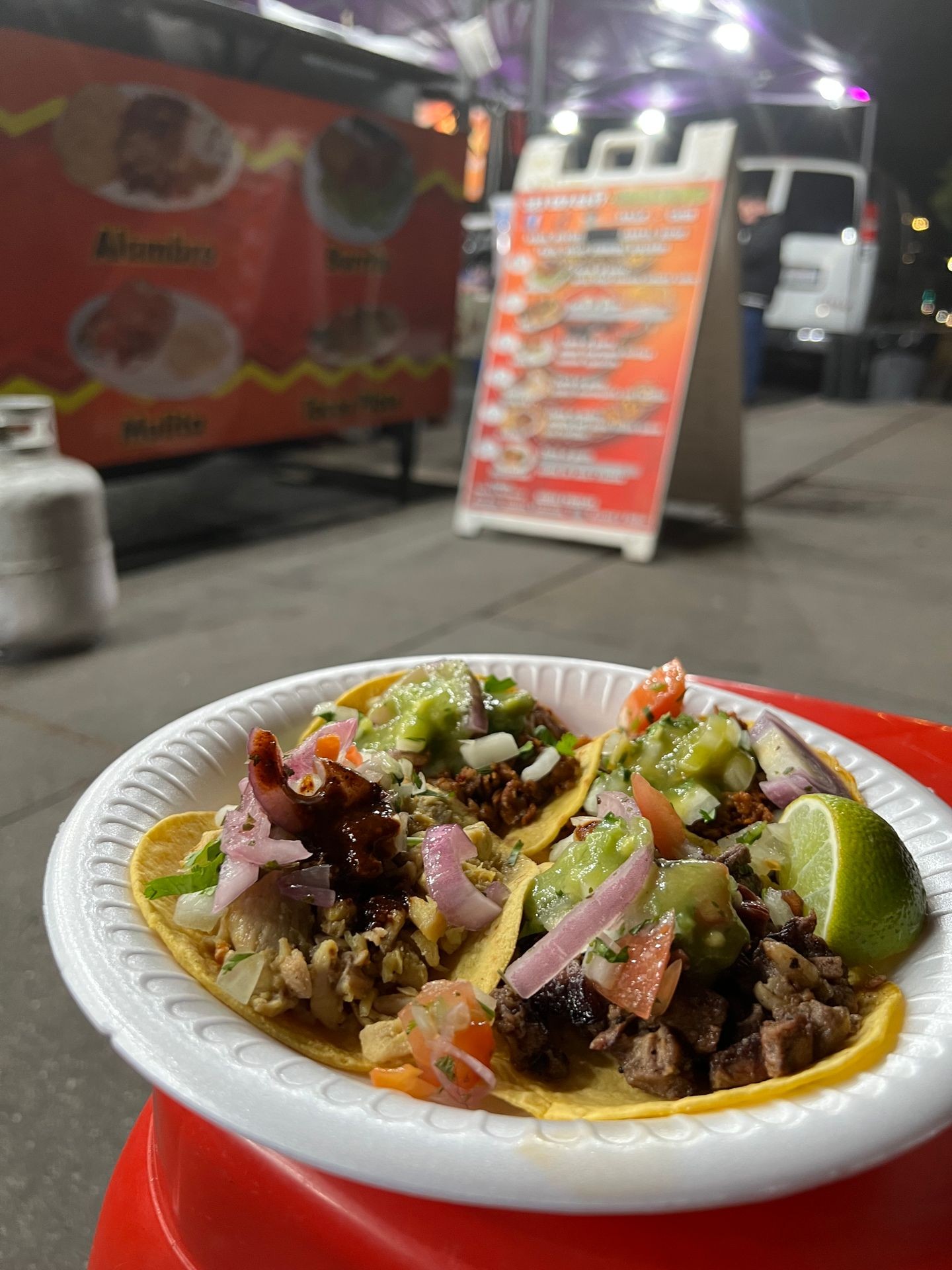 Three tacos on a paper plate with lime and toppings, street food stand in the background.