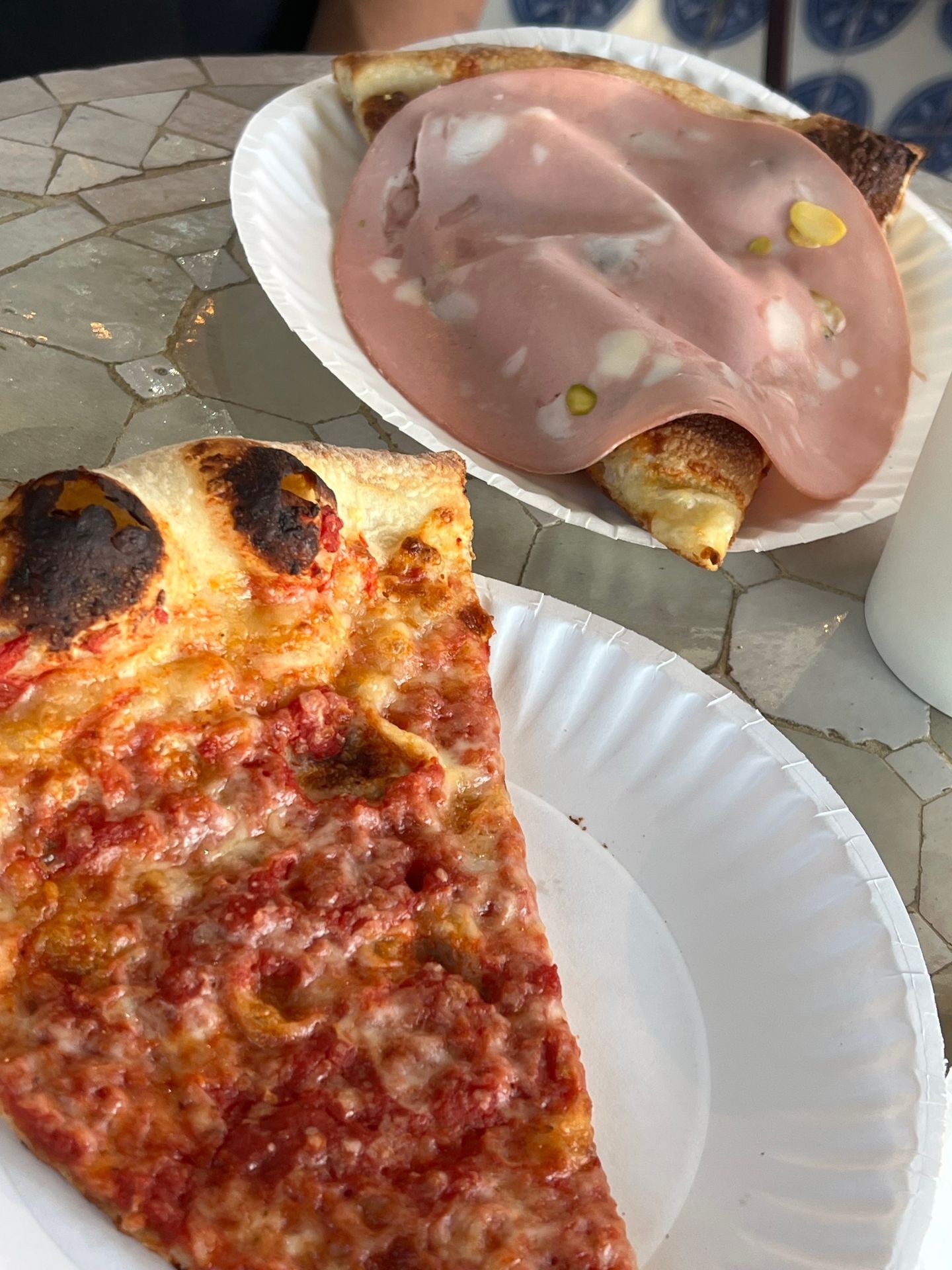 Burnt cheese pizza slice and mortadella pizza slice on paper plates.