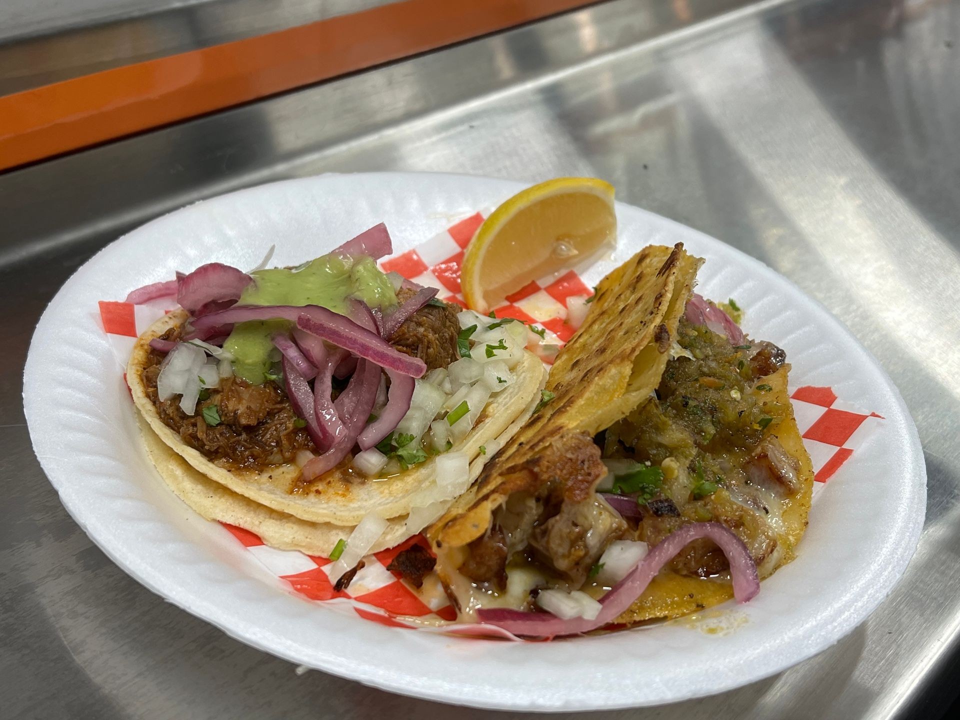 Two tacos with onions, cilantro, and green sauce on a paper plate with a lemon wedge.