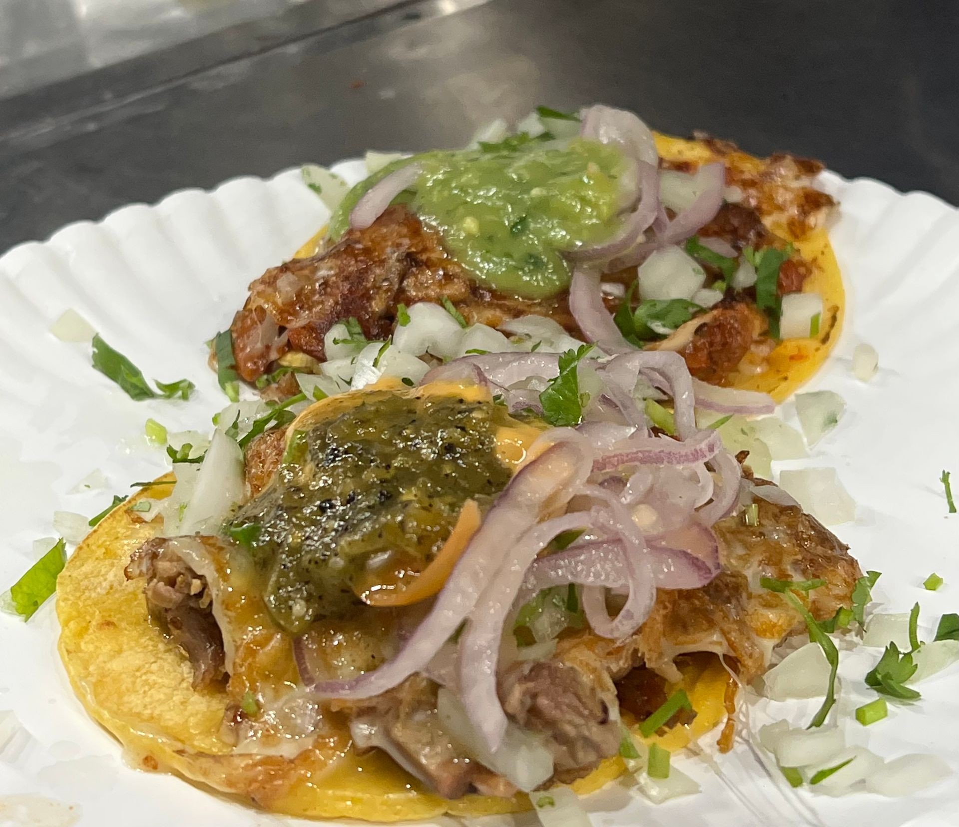 Close-up of two tacos topped with onions, cilantro, and green salsa on a paper plate.