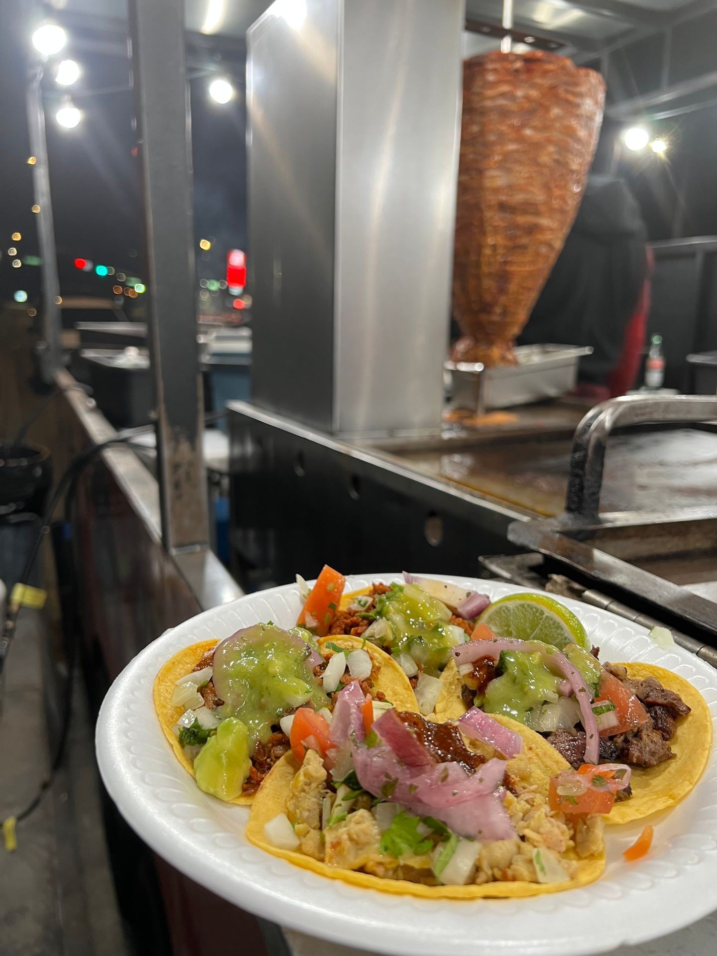 Street tacos with various toppings on a paper plate, near a vertical meat rotisserie at night.